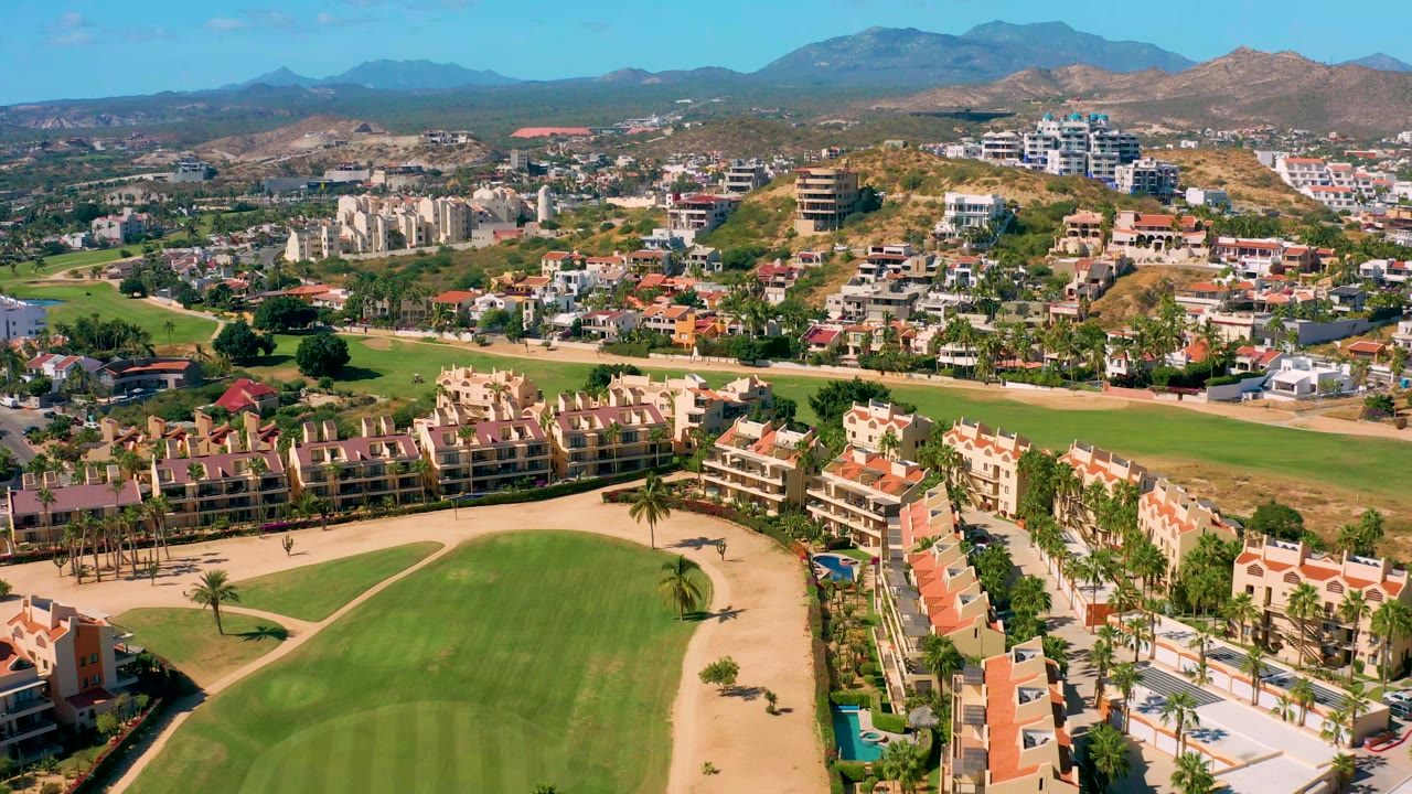 A wide San Jose del Cabo aerial shows the fairway and sand course edges below, clustered homes and apartment blocks beyond, a hillside development, mountain horizon, and no visible people.