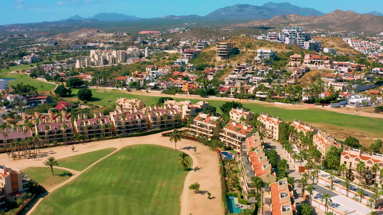 The aerial view over San Jose del Cabo holds on green course turf, tan paths, cream residential blocks with red roofs, hillside houses, hazy mountains, and no visible people.