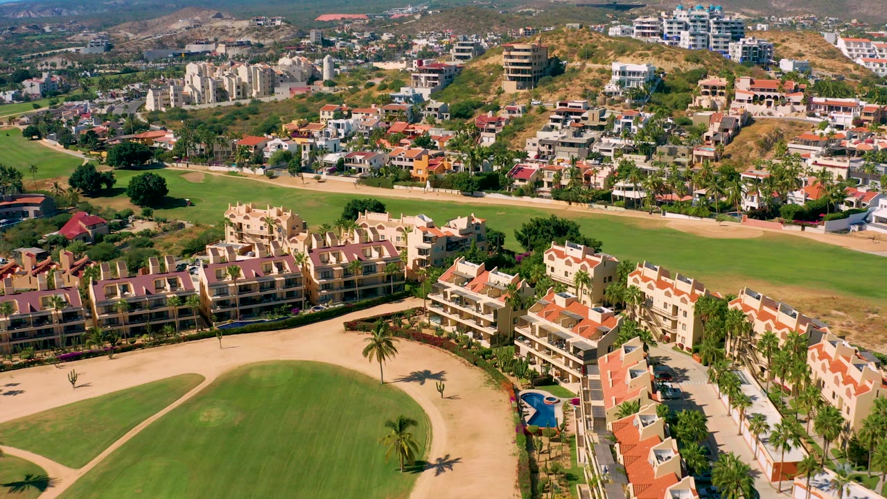 Aerial frame over San Jose del Cabo showing a curved green fairway, sandy bunker paths, red-roofed residential buildings, palm trees, hillside homes, and no visible people.