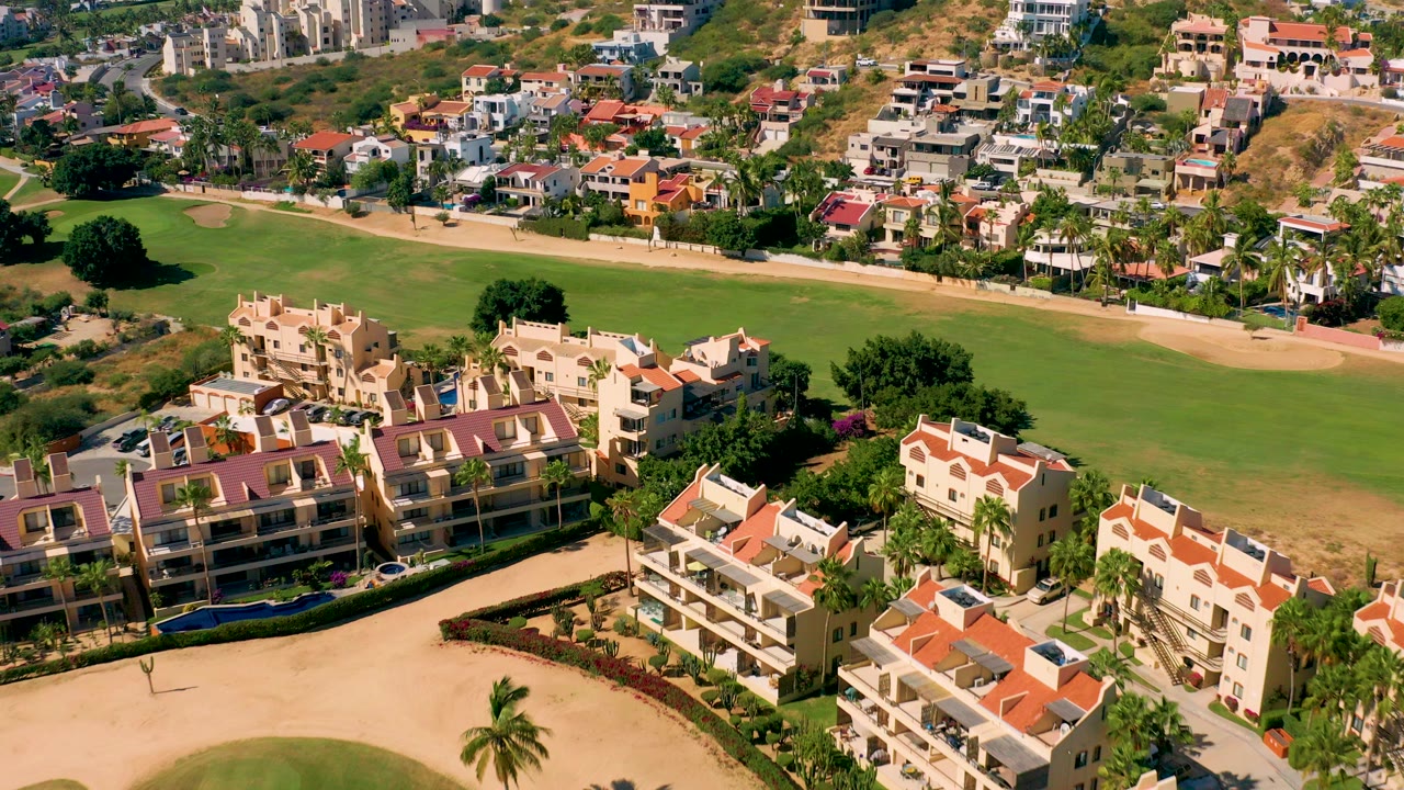 Aerial frame over San Jose del Cabo homes with red tile roofs, a green golf fairway behind them, sandy paths, palm trees, and no visible people.