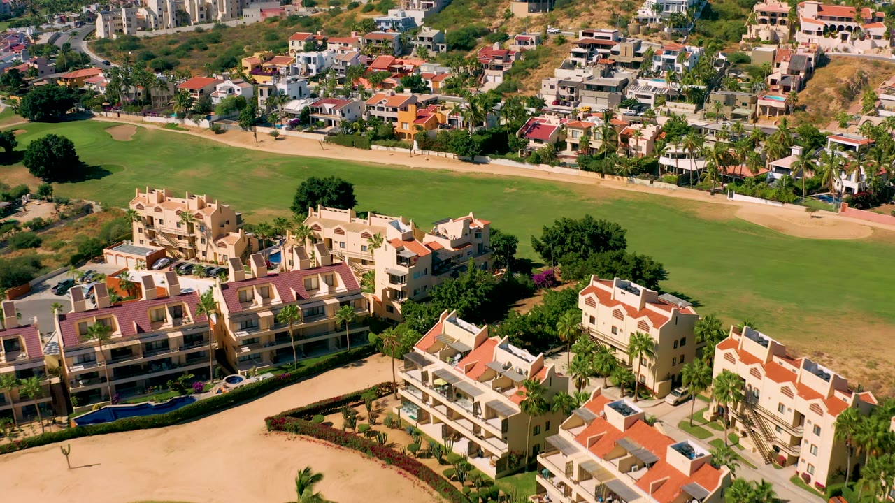 San Jose del Cabo aerial still with clustered red-roof residences at the lower edge, open golf course grass through the center, hillside houses beyond, and no visible people.