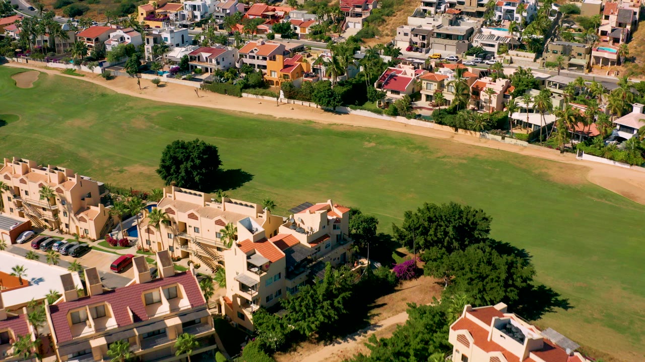 Aerial view in San Jose del Cabo showing cream homes with red roofs along a broad green field, a curved tan path, parked cars, palm trees, and no visible people.