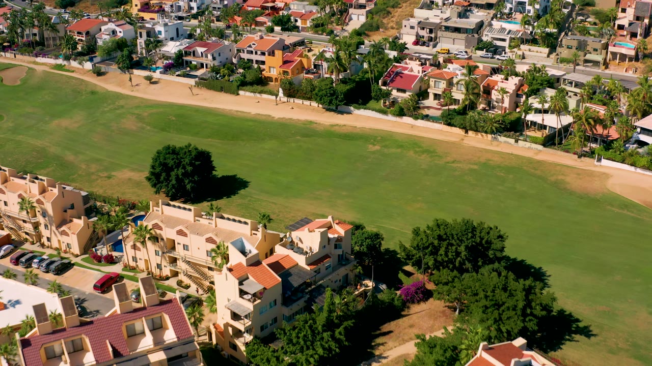 Aerial view in San Jose del Cabo with a large lawn filling the middle of frame, a tan path behind it, colorful homes in the background, foreground rooftops, and no visible people.