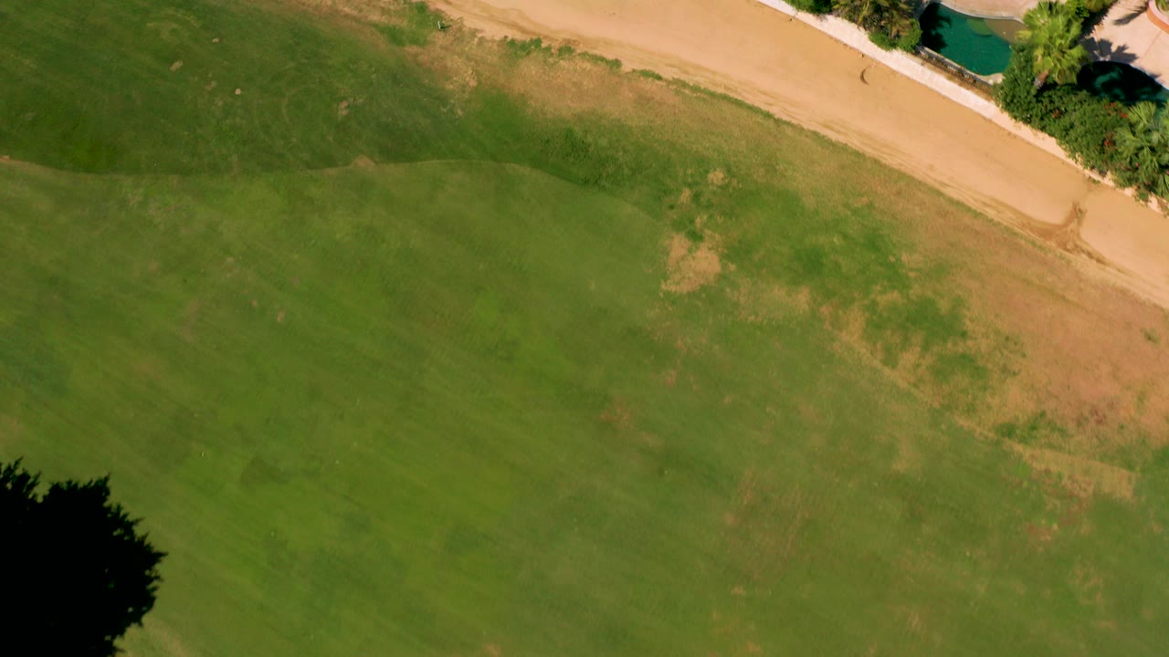 Overhead frame in San Jose del Cabo showing patchy green field below a tan sandy path, with tree shadow at the lower left and no visible people.