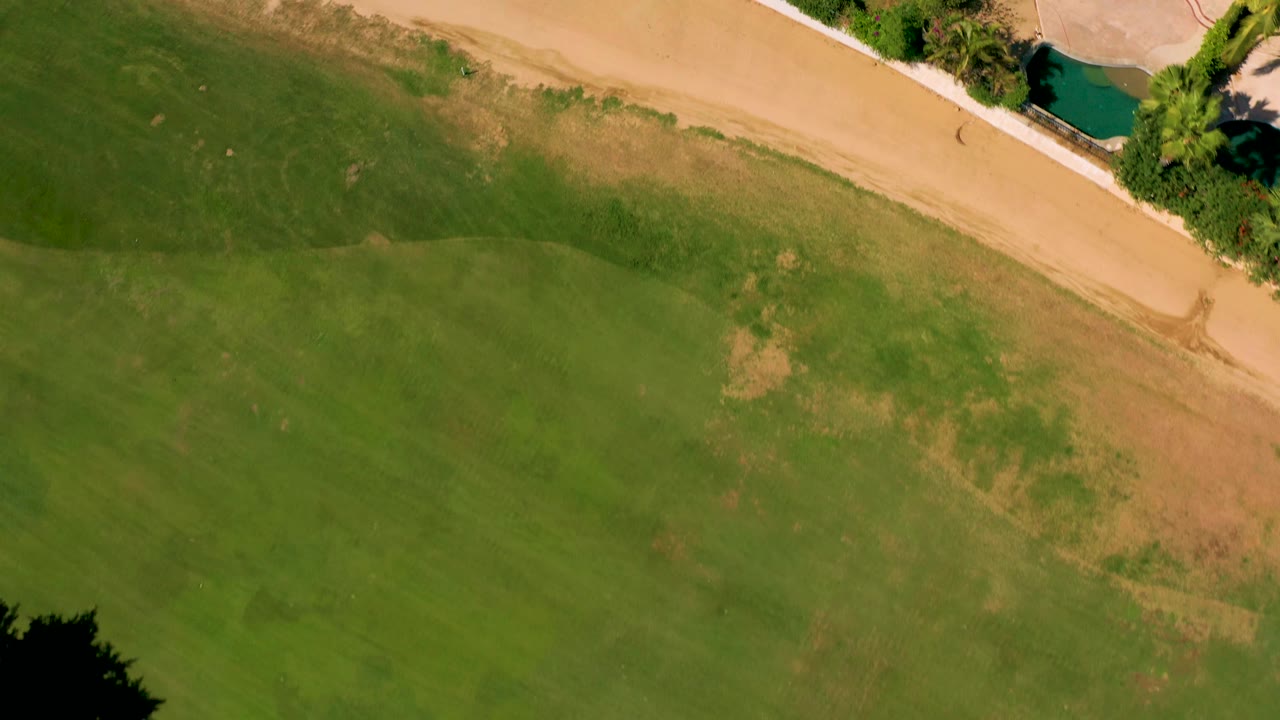 Top-down San Jose del Cabo frame showing grass below a sandy path, dense trees at the upper right, a turquoise pool, and no visible people.