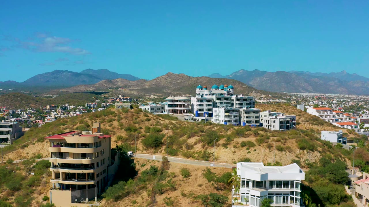 Opening frame of San Jose del Cabo hillside homes, dry scrub, a curving road, distant city blocks, mountain ridges, and no visible people.