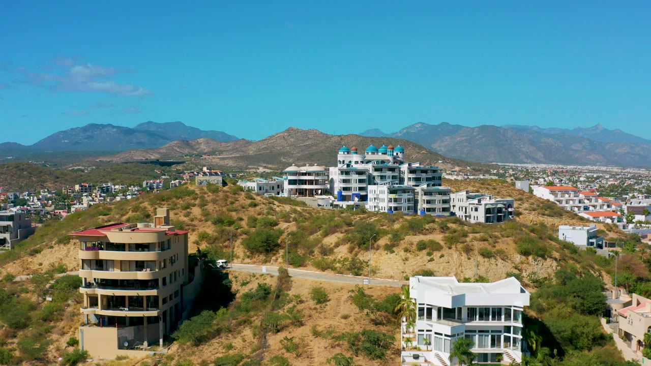 Elevated San Jose del Cabo view with dry vegetation across the hill, a road cutting along the slope, clustered modern residences, mountains, and no visible people.
