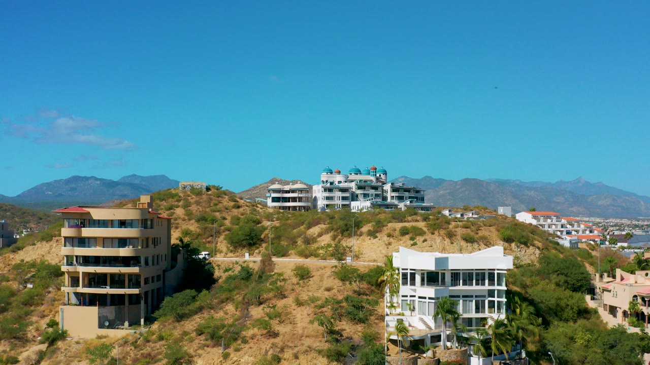 Wide drone view of hillside residential buildings in San Jose del Cabo with dry terrain, scattered greenery, distant mountains, and no visible people.