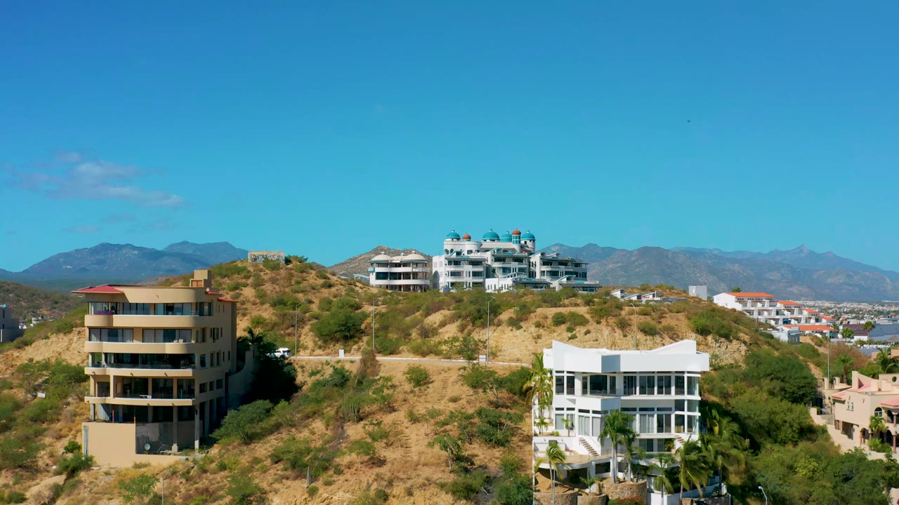 Drone frame over San Jose del Cabo hillside homes, with a beige building at left, a white building at lower right, dry slopes, and no visible people.