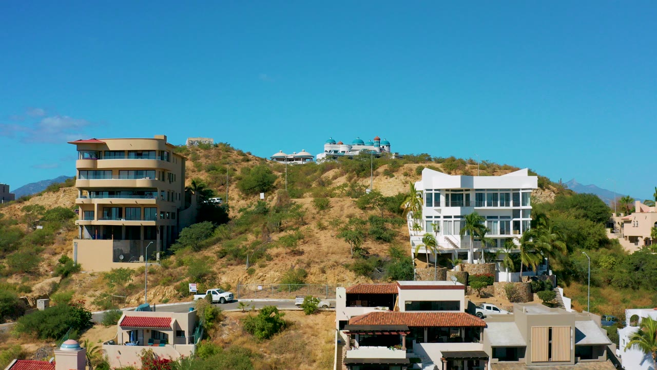 Opening frame of San Jose del Cabo hillside homes, a winding road, dry grass, shrubs, and a clear blue sky with no visible people.