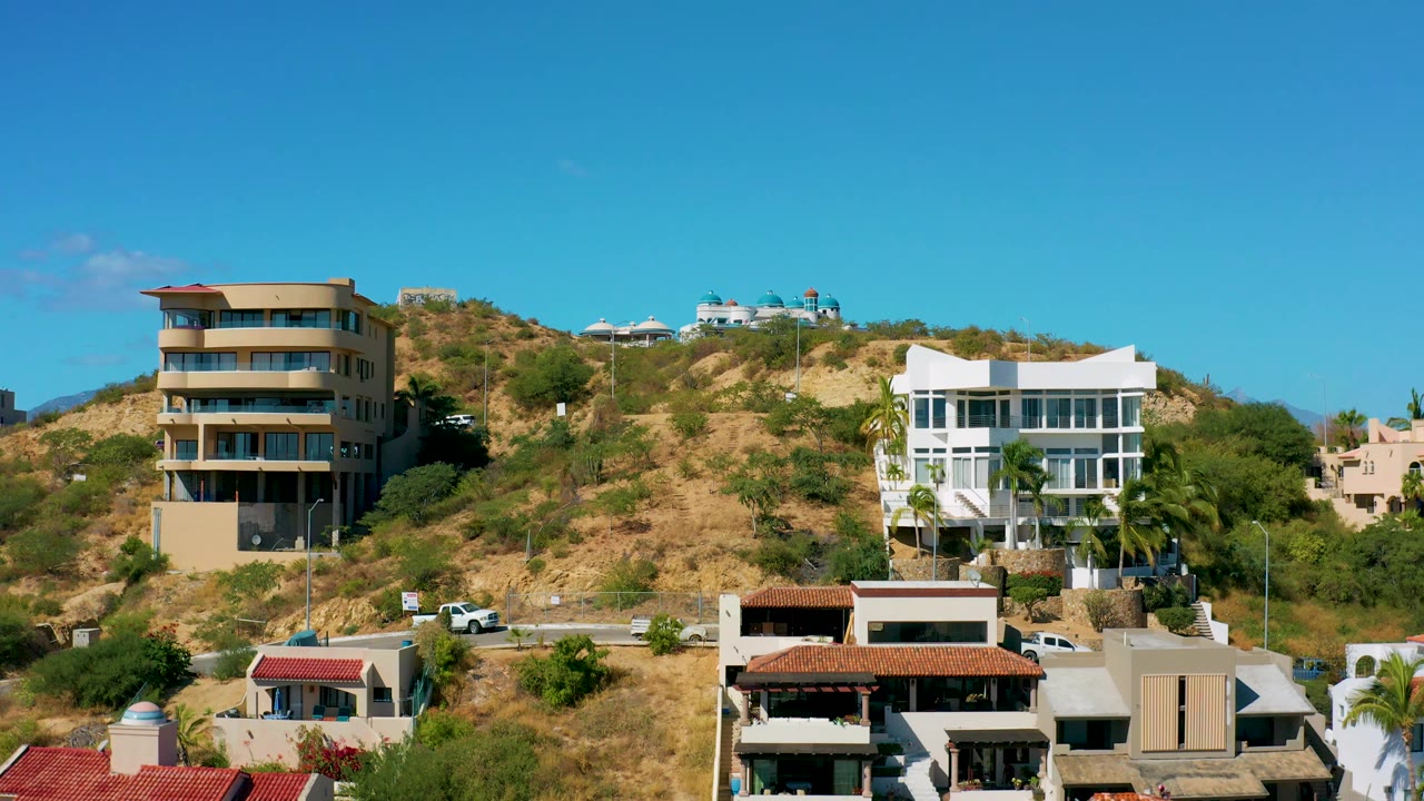 A San Jose del Cabo hillside view centers the dry slope between a tan building and a white modern house, with vehicles on the road and no visible people.