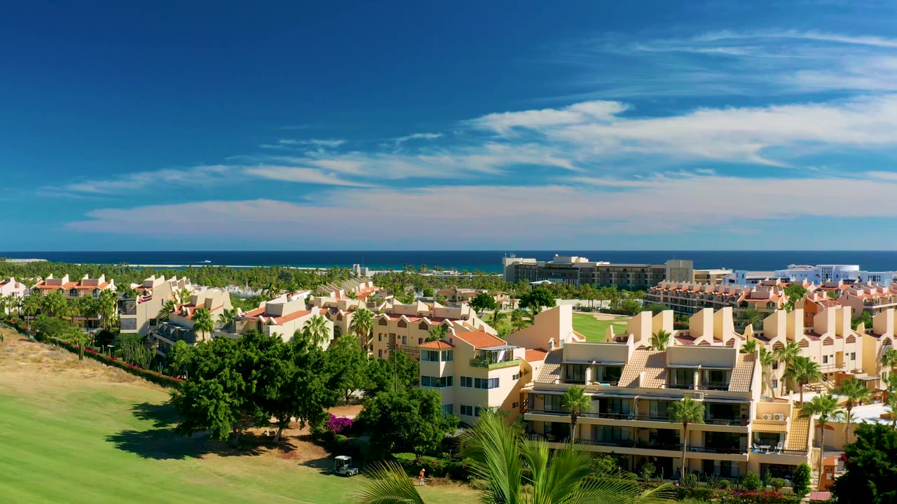 At 0:01, the San Jose del Cabo resort view holds steady with palms in front, beige buildings at center, a dark ocean line beyond, and no visible people.