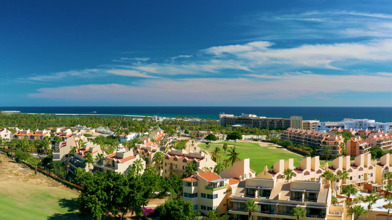 Opening aerial frame over San Jose Del Cabo residential buildings and a green golf course with the ocean horizon beyond, a small white boat offshore, and no visible people.