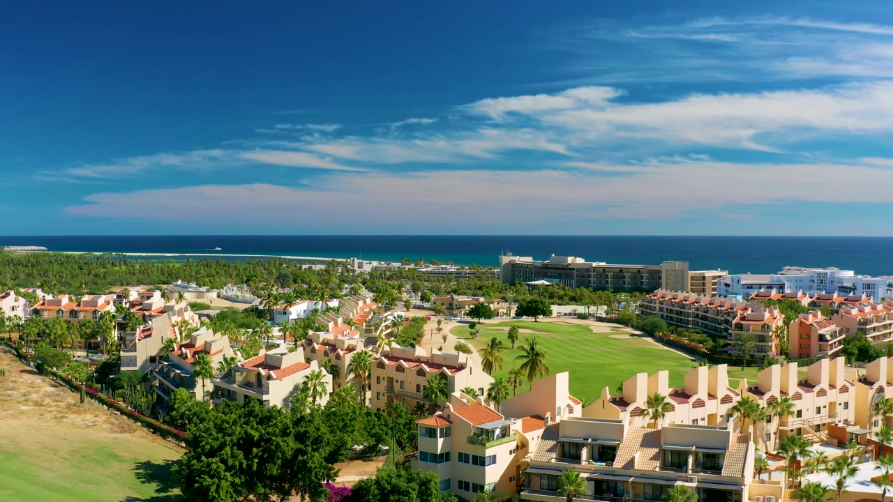 San Jose Del Cabo aerial frame showing more of the residential blocks around the golf course, with palm-lined greenery, ocean surf color near shore, a small white boat, and no visible people.