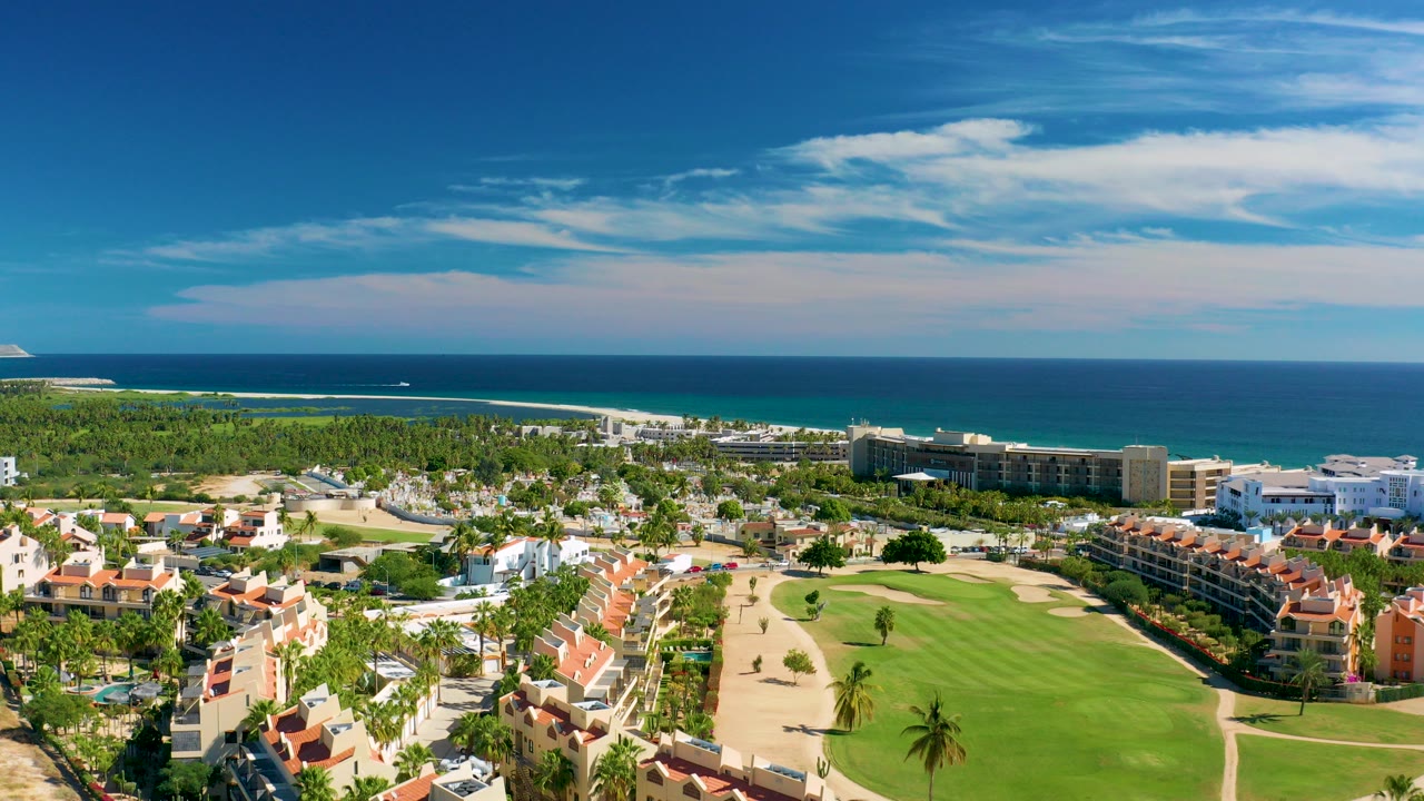 Wide aerial frame over San Jose Del Cabo resort buildings and a golf course, with beach and turquoise ocean beyond and no visible people.