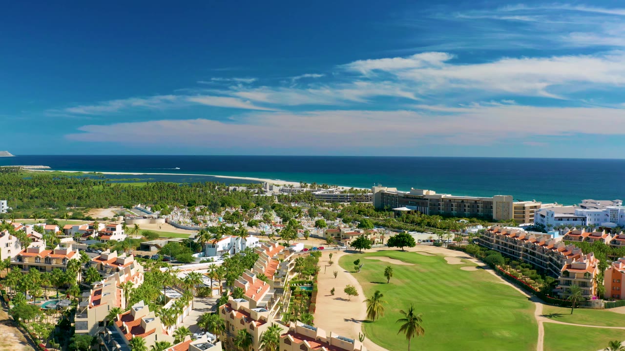 San Jose Del Cabo coastal resort scene with the fairway centered, palm trees and red-roof residences below, and a clear ocean horizon in the background, no visible people.