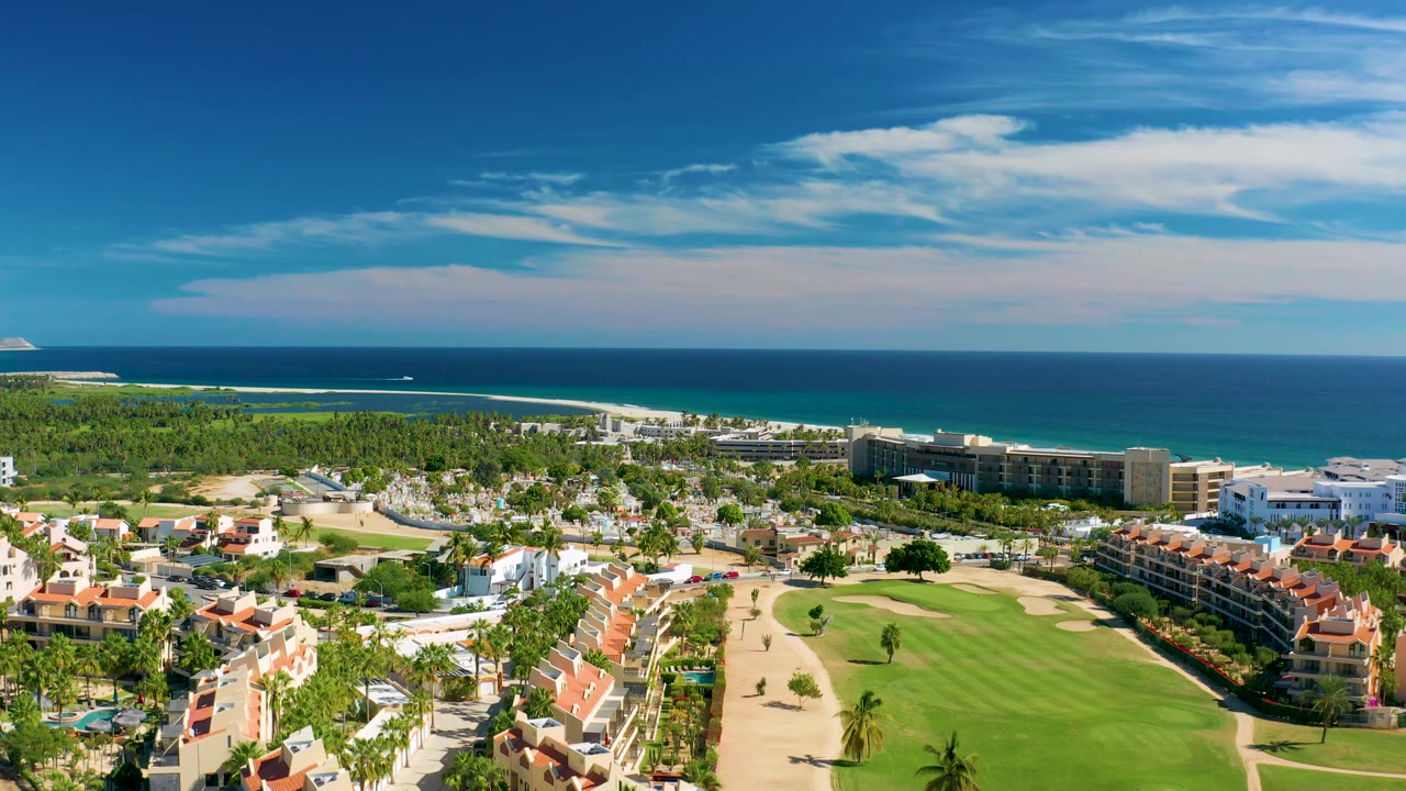 Aerial view in San Jose Del Cabo showing red tile rooftops in the foreground, a green golf fairway and sand areas in the middle, and ocean water behind, no visible people.