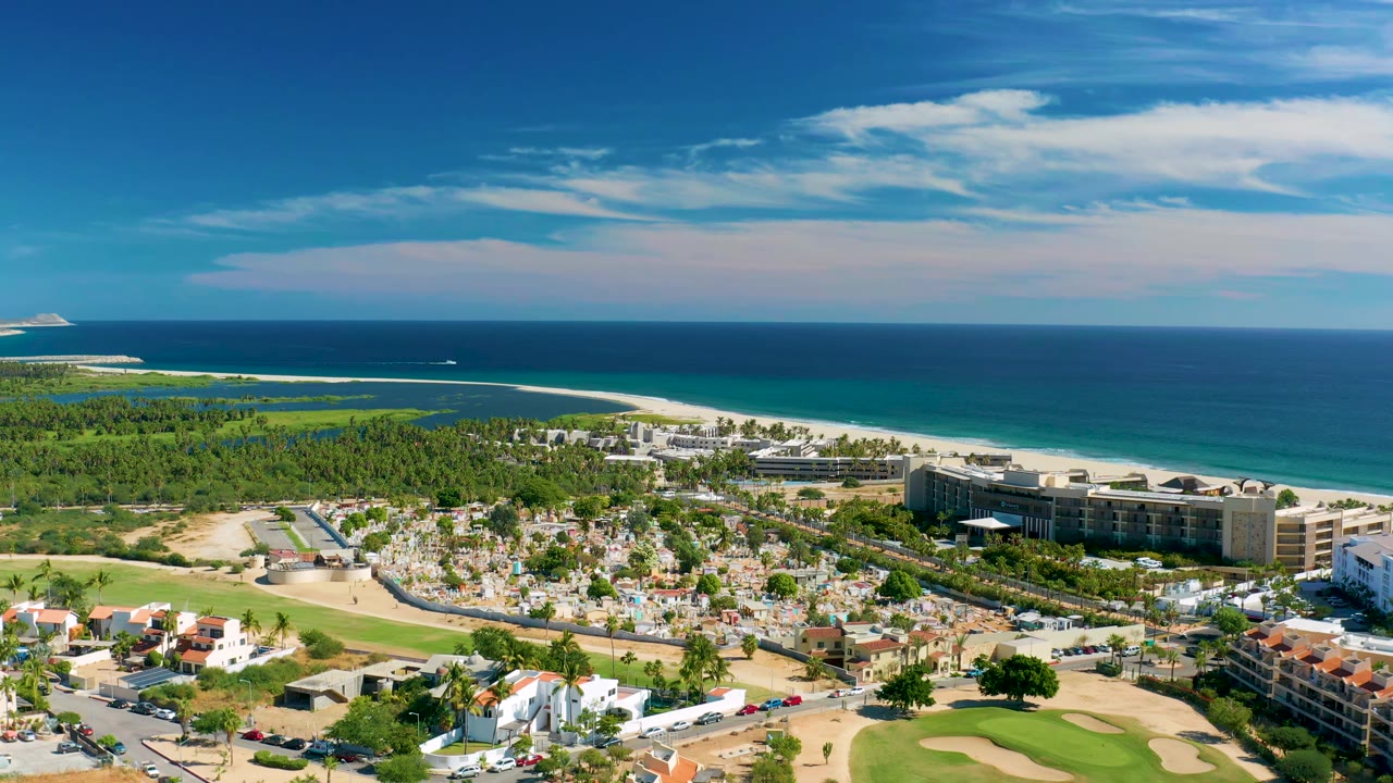 Wide aerial frame in San Jose Del Cabo showing a resort complex beside the beach, small vehicles on roads, ocean bands, green landscape, and no visible people.