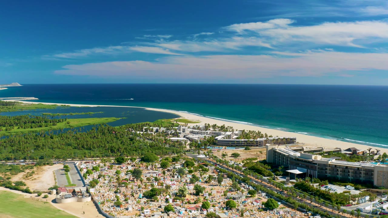 Drone-like coastal view with ocean horizon, resort buildings, beach, road, and foreground plot grid.