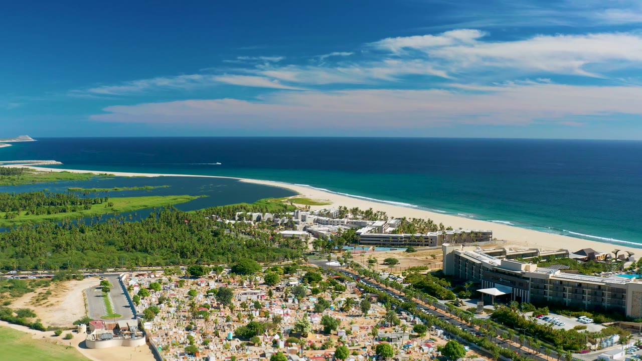 Wide San Jose del Cabo coastal frame showing turquoise water, white surf line, resort buildings near the beach, green inland vegetation, roads, and no visible people.
