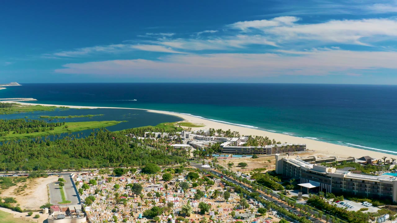 Coastal aerial with beach, ocean, resort buildings, green wetlands, and foreground roads.
