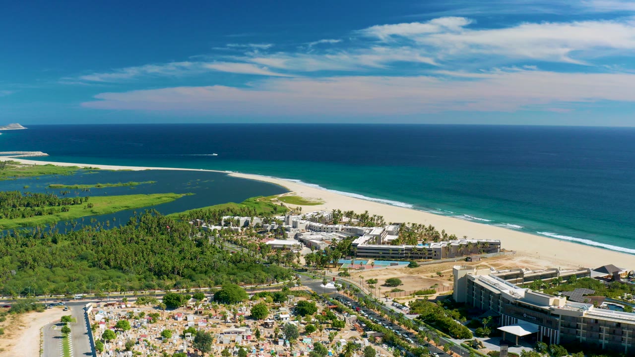 Opening aerial frame over San Jose Del Cabo with resort buildings, green vegetation, white sand, turquoise water, deep blue ocean, a distant boat, and no visible people.