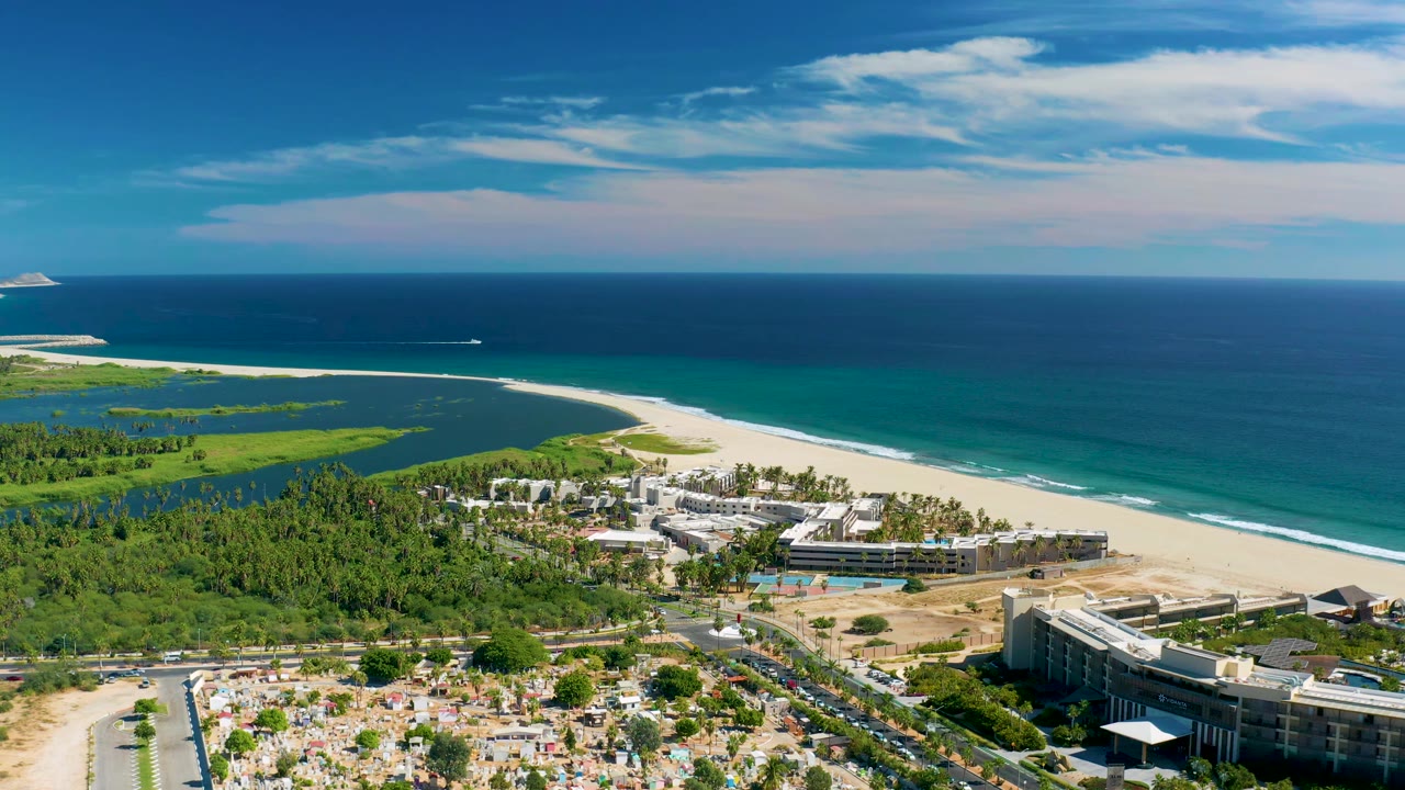 Aerial frame in San Jose Del Cabo showing the resort complex lower in frame, a road and parking area at the bottom, green land to the left, beach and ocean beyond, and no visible people.