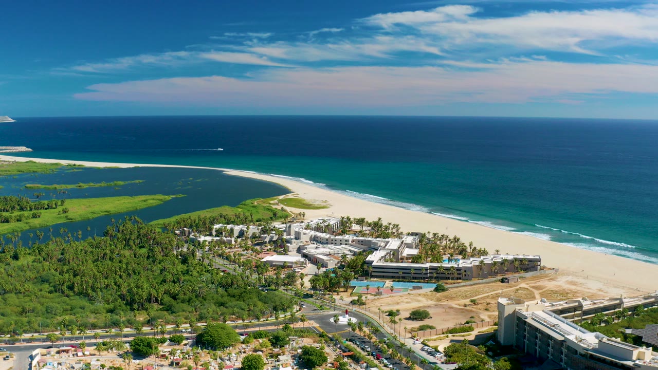 Aerial view in San Jose Del Cabo showing resort buildings, road traffic, green vegetation, lagoon water, sandy beach, ocean waves, and no visible people.