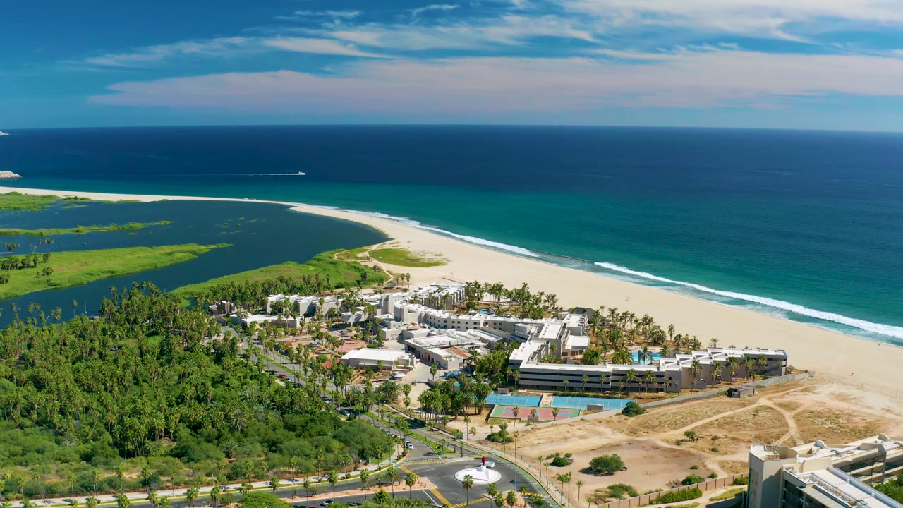 San Jose del Cabo aerial frame with the lagoon on the left, a pale beach curving along the ocean, coastal buildings below, and no visible people at 0:01.