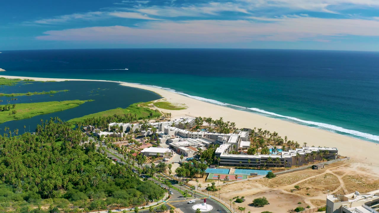 San Jose del Cabo aerial still with the lagoon and greenery at left, beach and surf across the middle, coastal buildings below, and no visible people at 0:03.