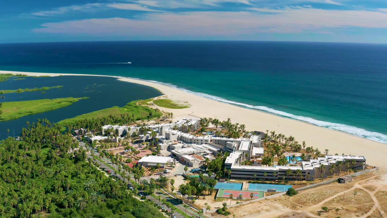 Elevated San Jose Del Cabo coastline view showing the resort lower in frame, lagoon to the left, a long white beach through the center, and dark blue ocean beyond, with no visible people.