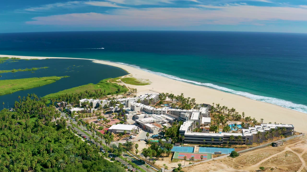 Aerial frame from San Jose Del Cabo with resort roofs and pool areas at the bottom, green vegetation at left, a sweeping sand beach, lagoon edge, and blue ocean, with no visible people.