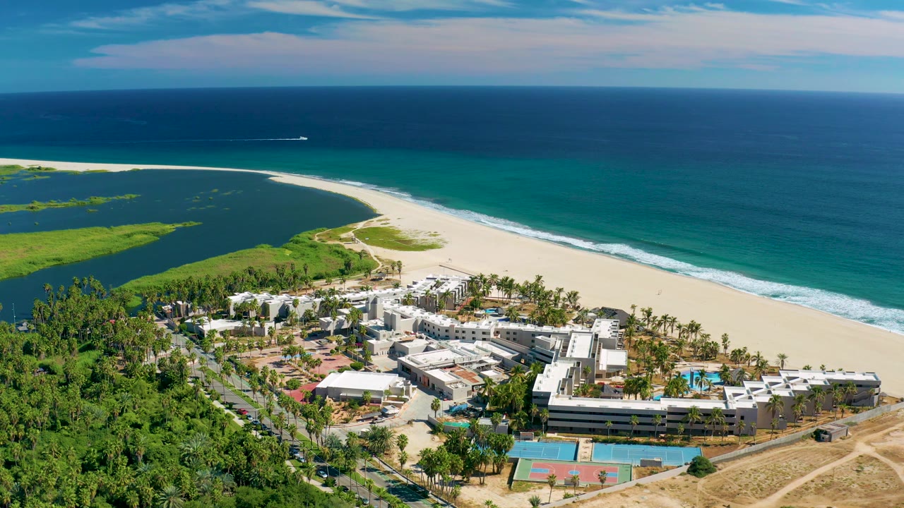 San Jose Del Cabo aerial view with the lagoon and green shoreline on the left, resort buildings and palms below, white sand in the middle, and a blue ocean horizon, with no visible people.