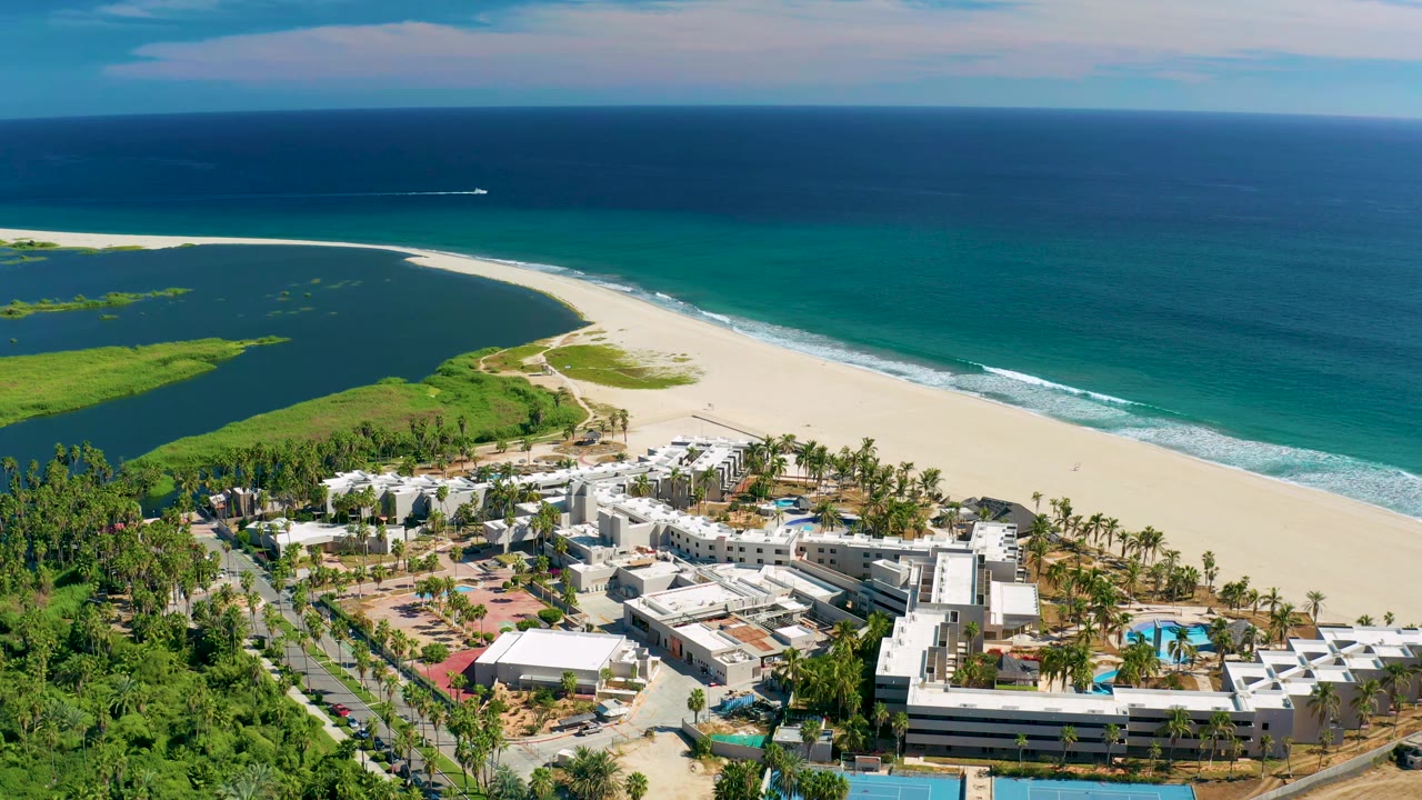 San Jose Del Cabo aerial frame showing white resort roofs in the foreground, a curved sandy beach, green inlet water, blue ocean, and no visible people.