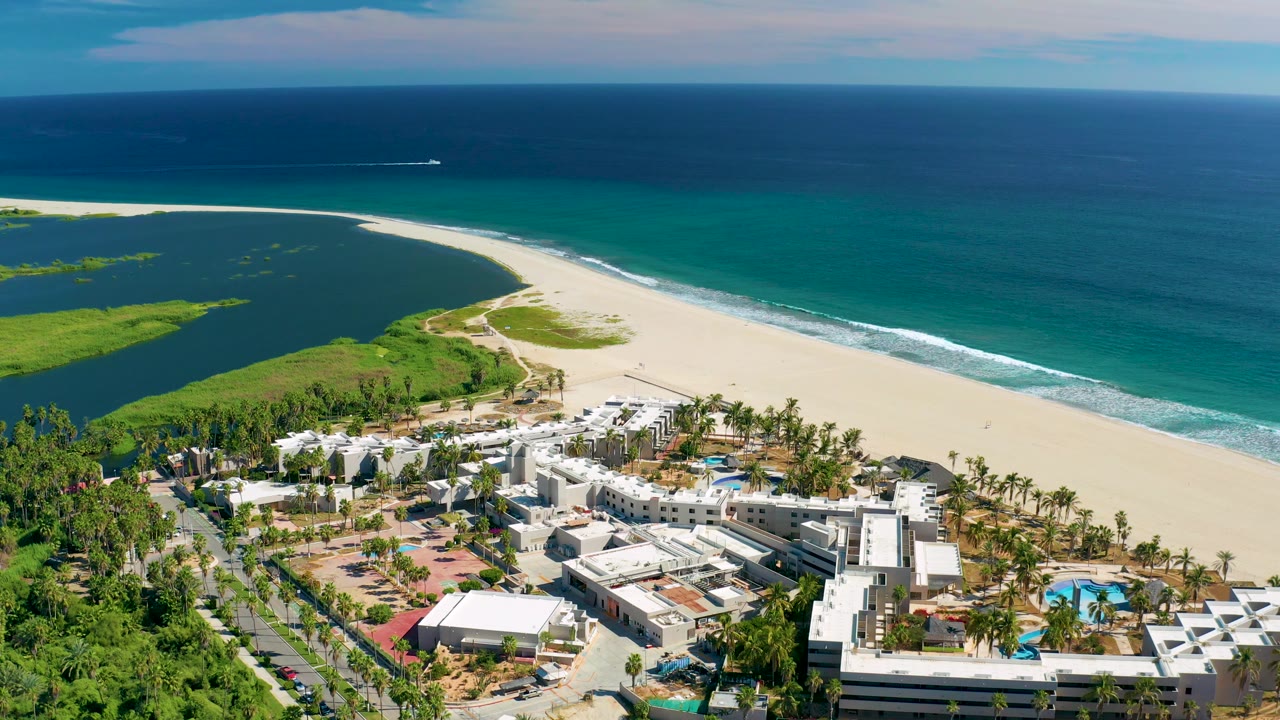 Aerial San Jose Del Cabo view with the resort complex lower right, green vegetation lower left, pale beach across the middle, blue ocean beyond, and no visible people.