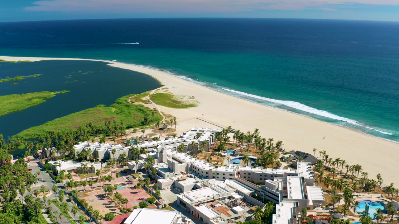 Opening aerial frame over San Jose del Cabo resort buildings, palm trees, pale beach, green lagoon, and blue ocean, with no visible people.
