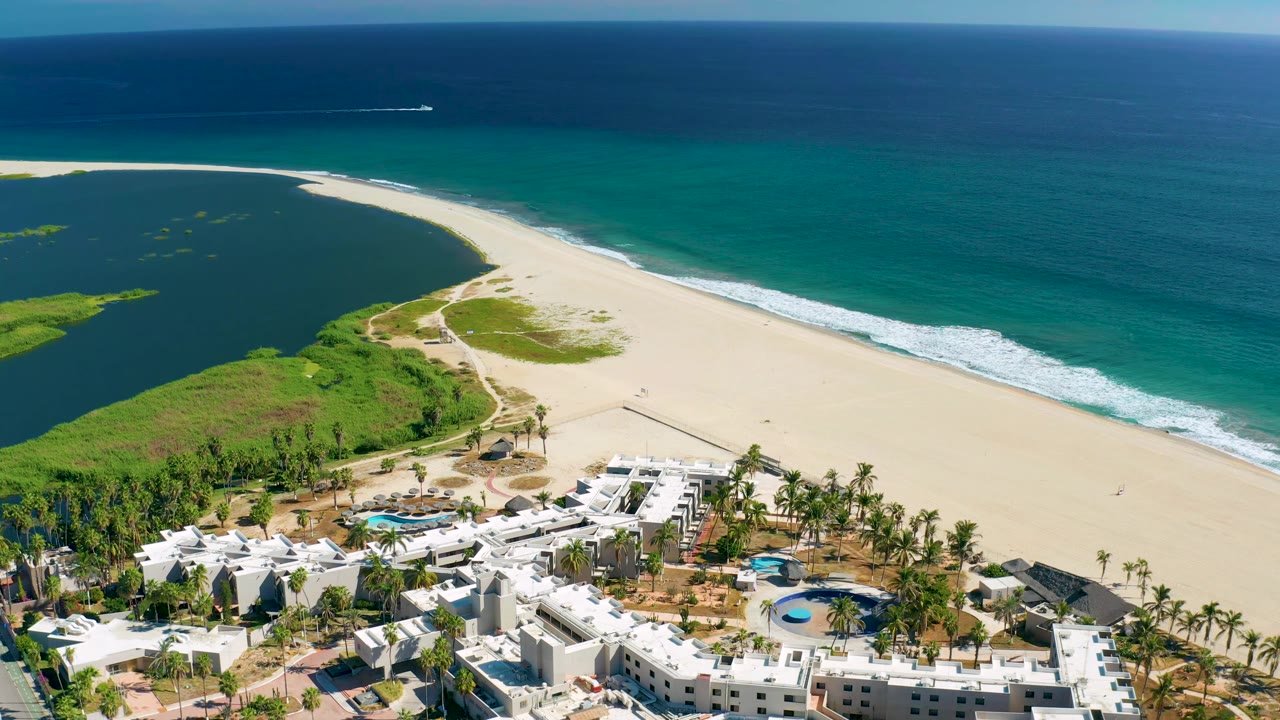 Aerial view in San Jose Del Cabo with the resort slightly lower in frame, green vegetation along the sand, turquoise surf, open ocean, and no visible people.