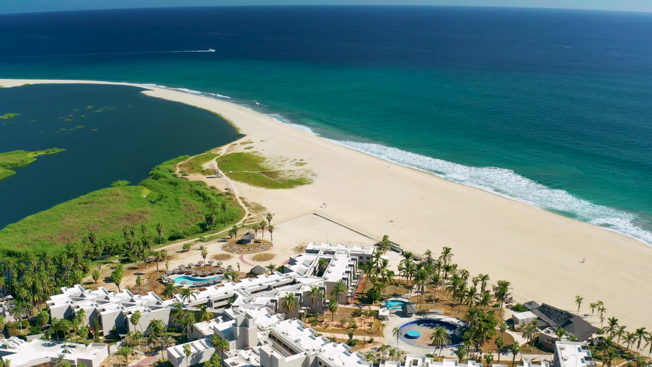 Mid-clip aerial over San Jose Del Cabo showing resort rooftops, palm rows, a broad sandy beach, green wetland-like vegetation, turquoise water, and no visible people.