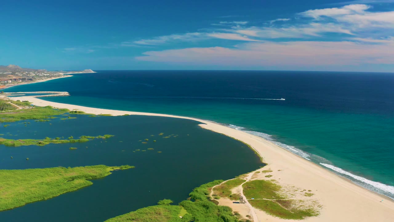 Opening aerial frame over San Jose del Cabo with green lagoon in the foreground, pale beach curving beside turquoise water, a small boat offshore, and no visible people.