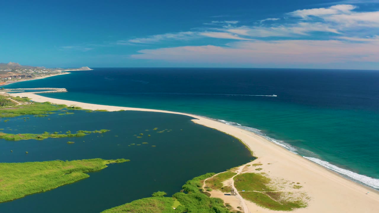 Mid-clip aerial frame over San Jose del Cabo with the lagoon occupying the lower left, the beach cutting across the frame, the boat farther along offshore, and no visible people.