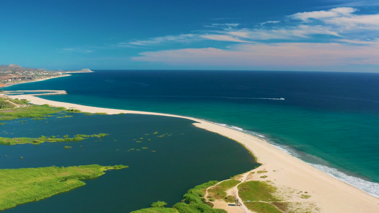 Aerial frame over San Jose del Cabo showing a curved sandy beach separating green lagoon water from turquoise ocean, with no visible people.