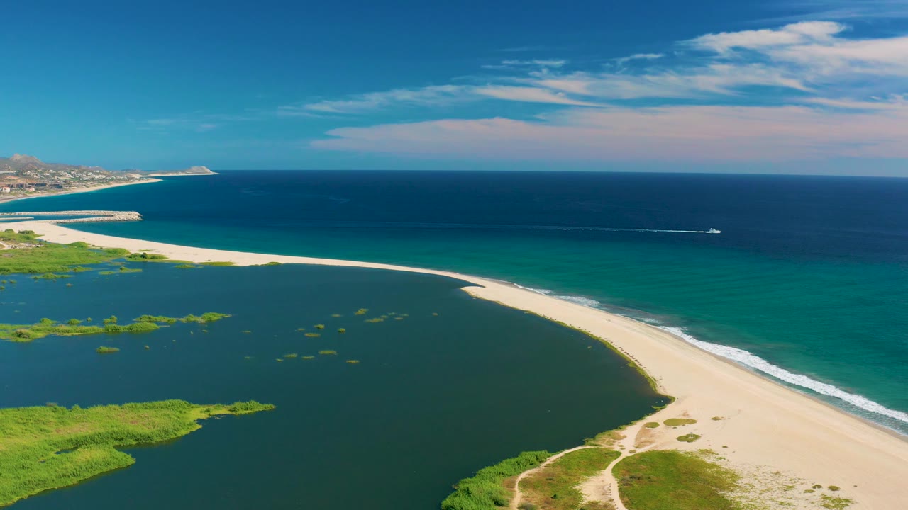 Wide aerial frame over San Jose Del Cabo with lagoon water across the foreground, a narrow sandy beach dividing the frame, ocean and horizon beyond, a boat wake to the right, and no visible people.