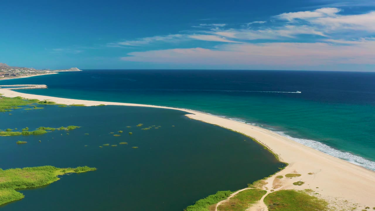 Aerial frame in San Jose del Cabo with the curved sandbar cutting between dark lagoon water and turquoise ocean, while the white boat sits farther right offshore and no people are visible.