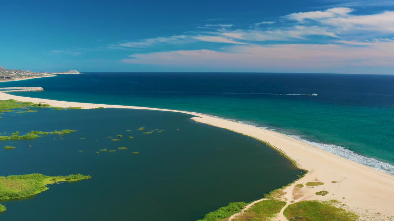 Aerial view over San Jose del Cabo lagoon and beach, with green vegetation patches at the lower edge, open ocean beyond the surf, a small boat offshore, and no visible people.