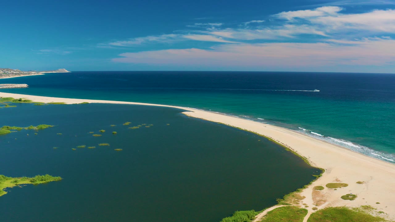 Aerial frame from San Jose Del Cabo shows a pale sandbar dividing dark lagoon water from blue ocean, with a small boat and wake offshore and no visible people.