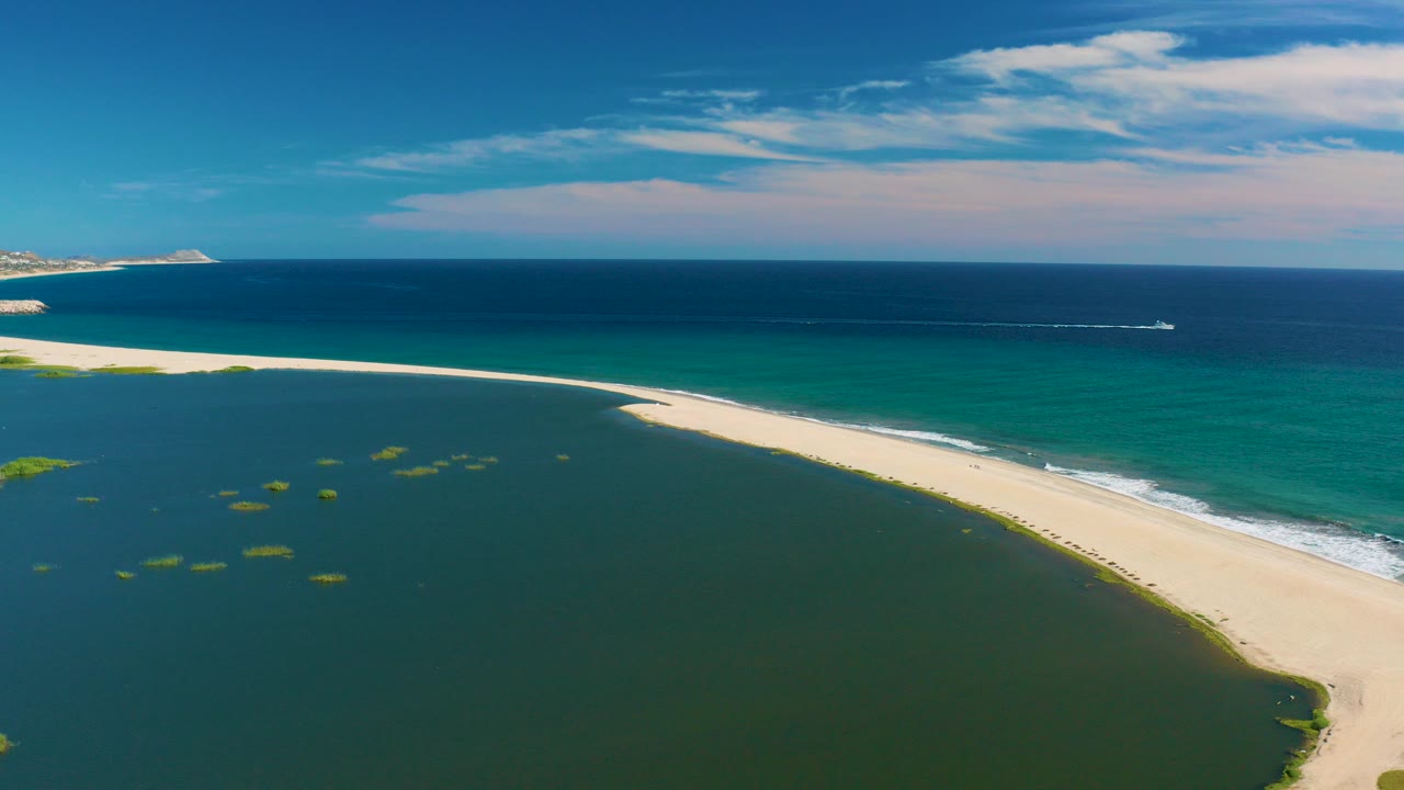 Aerial view in San Jose del Cabo, Baja California Sur, showing a curved pale sand beach between green lagoon water and blue ocean with a small boat wake offshore and no visible people.