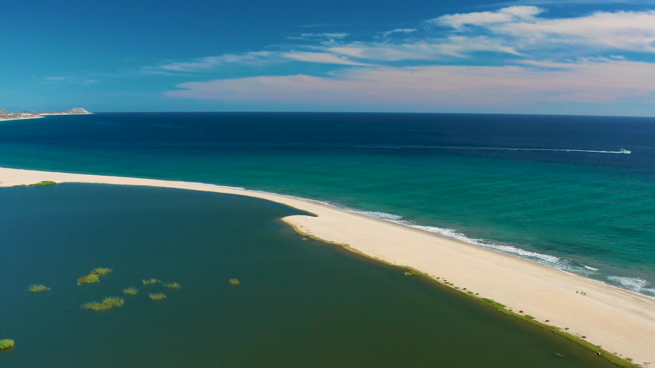 San Jose Del Cabo drone-style view with lagoon filling the lower left, a bright beach strip across the middle, deep blue ocean behind it, a small white wake to the right, and no visible people.
