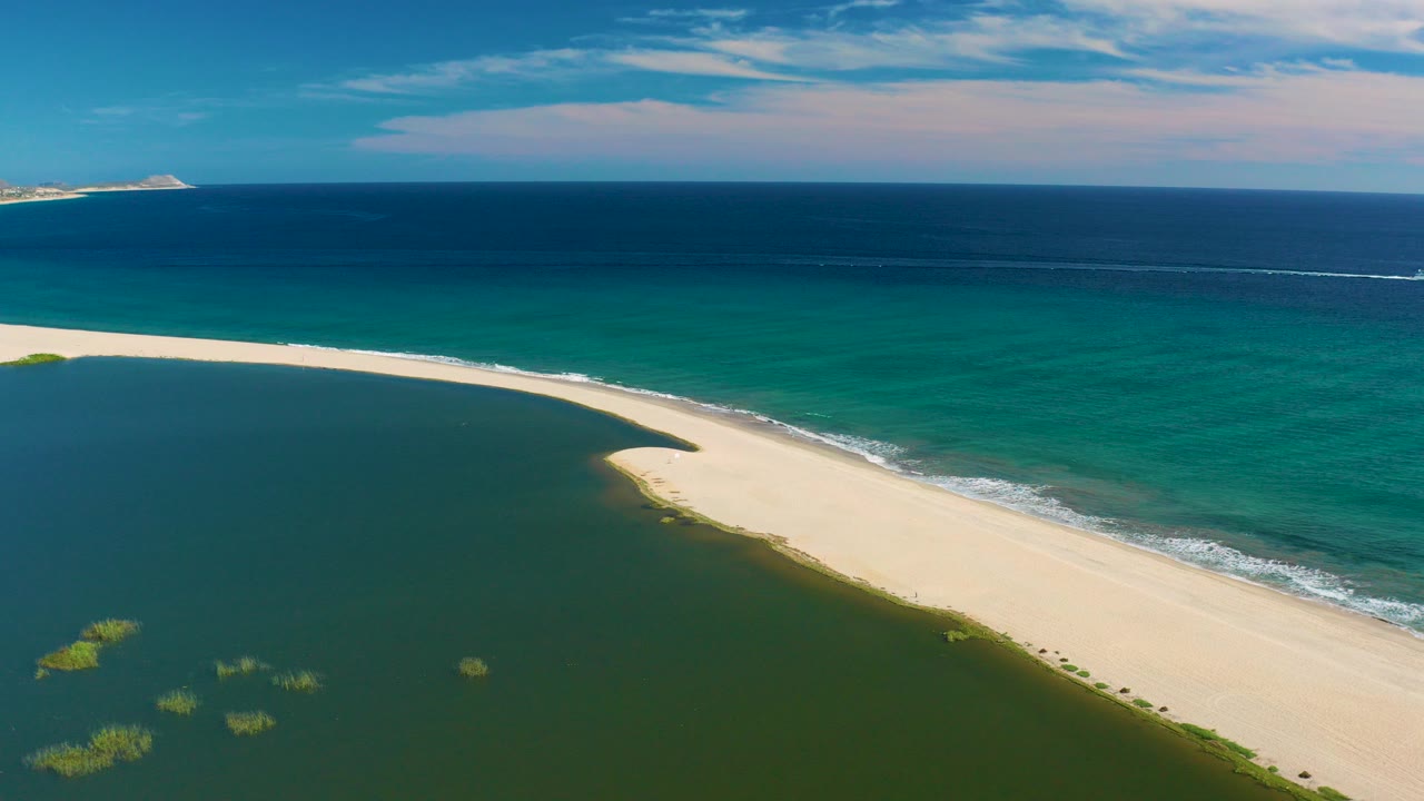 Aerial frame over San Jose Del Cabo showing a pale sandy spit separating green lagoon water from turquoise and deep blue ocean, with no visible people.