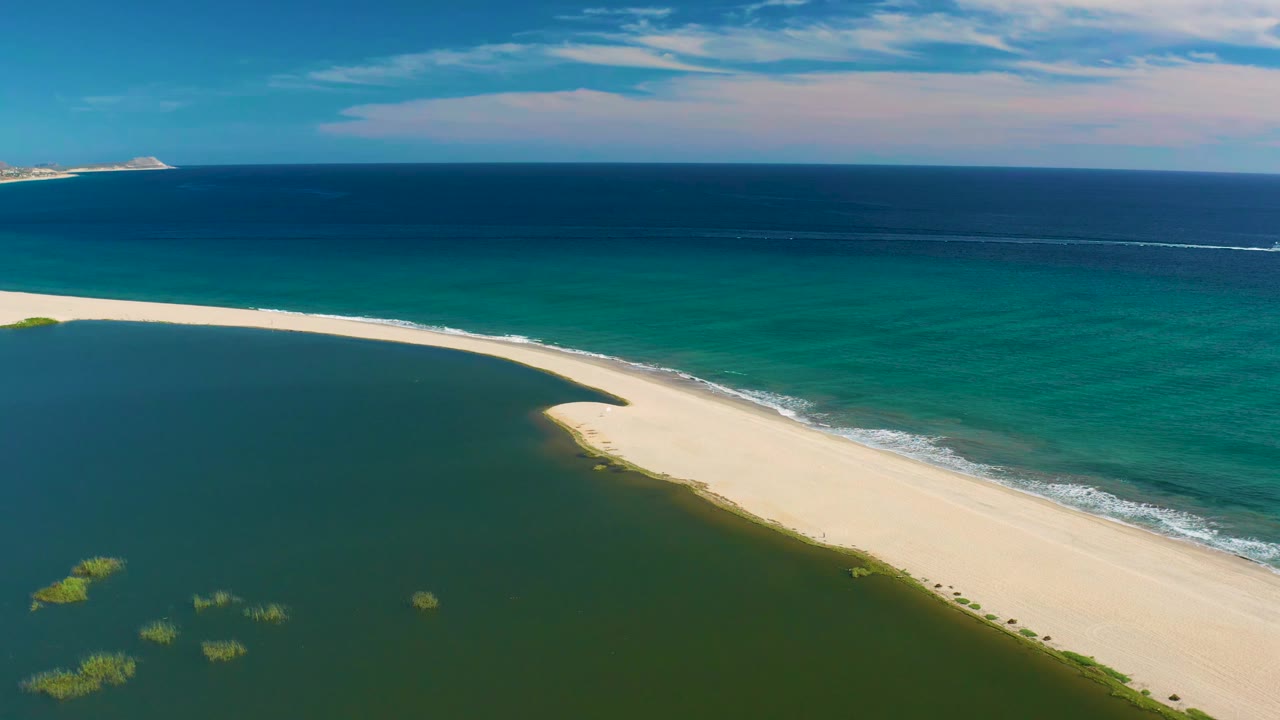Aerial view in San Jose Del Cabo with the curving sandbar shifted slightly right, green lagoon water below, ocean bands beyond, and no visible people.