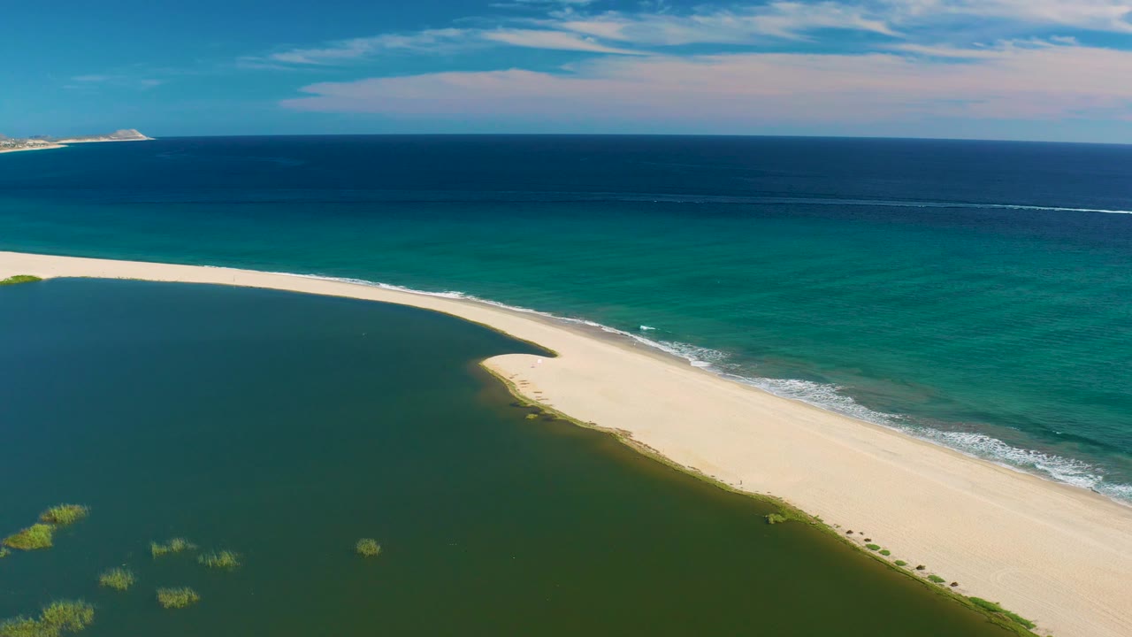 San Jose Del Cabo aerial frame showing the narrow sand spit, a darker lagoon on one side, small white waves on the ocean side, and no visible people.